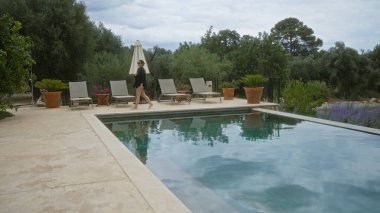 İspanya, Mallorca 'da yüzme havuzunun yanında yürüyen bir kadın. Etrafı yeşillik ve şezlonglarla çevrili. Bulutlu gökyüzünün altında.