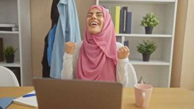 Ev ofis ortamında başarıyı kutlayan tesettürlü neşeli bir kadın, masada bilgisayar ve bardak..