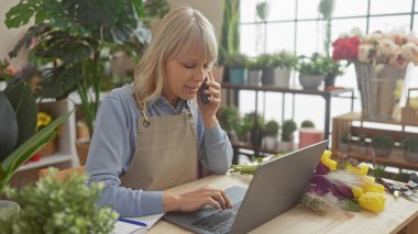 Önlüklü sarışın bir kadın dizüstü bilgisayar kullanıyor ve çiçeklerle çevrili bir çiçekçide telefonla konuşuyor.