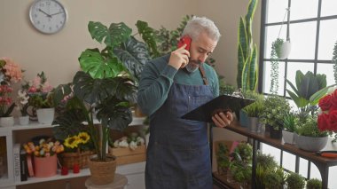 Önlüklü sakallı adam çiçekçi dükkanında telefon ve pano ile birden fazla iş yapıyor..