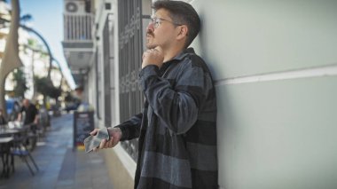 Yakışıklı, gözlüklü ve bıyıklı genç bir adam canlı bir şehir caddesinde telefon tutuyor..