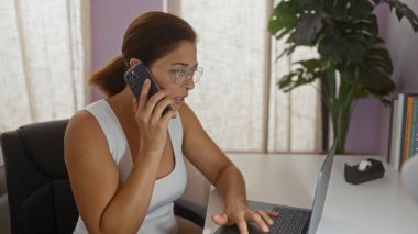 Ofis ortamında dizüstü bilgisayar kullanırken telefonla konuşan, odaklanmış ifade ve esmer saçlar sergileyen, arka planda iç mekan bitkileriyle ofis sandalyesinde oturan bir kadın.