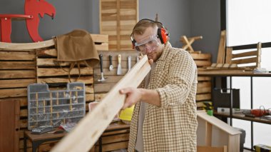 Sakallı genç bir adam, iyi organize edilmiş bir marangozluk atölyesinde, koruyucu gözlük ve kulaklık takan ahşap bir kalası inceliyor..