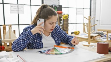 Hispanic woman multitasking on phone in a woodworking studio with color swatches and tools. clipart