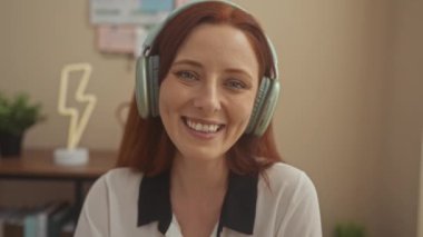 Kırmızı saçlı, gülümseyen genç bir kadın. Oturma odasında kulaklık takıyor. Dekoratif yıldırım lambası ve bitkileri var..