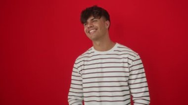 Yüzünde doğal bir gülümsemeyle kenarda duran çizgili tişört giyen genç İspanyol bir adam, kendine güvenen ve gülen, kırmızı bir arka planda izole bir halde.