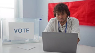 Ciddi bir ifadeye sahip genç İspanyol bir adam, Polonyalı bir seçim okulunda bilgisayar ve kulaklık kullanırken kameraya rahatça bakıyor.