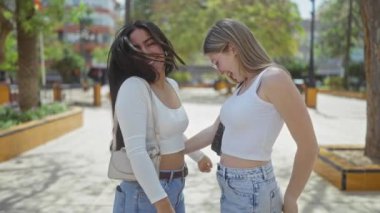 Güneşli bir şehir parkında ağaçlarla gülüp el ele tutuşan iki kadın.