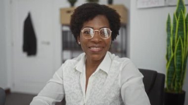 Ofiste gülümseyen gözlüklü ve arka planda bitki olan beyaz gömlekli bir kadın.