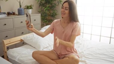 Kadın, arka planda bitki ve şifoniyerle rahat bir yatak odasında otururken akıllı telefon kullanarak video konferansı yapıyor.