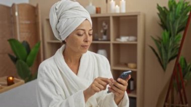 Bir kadın spa odasında mumlarla dinleniyor, telefona bakıyor, kafasına bornoz ve havlu giyiyor, sakin bir ortam, sağlık merkezi tesisi.