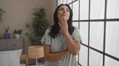 Mature woman looking thoughtfully out the window in a cozy bedroom setting with plants and soft lighting, wearing casual clothing and embracing introspective mood