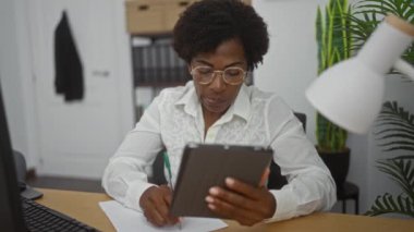 Orta yaşlı, Afro-Amerikalı bir kadın ofiste tablet okurken bir masada not alıyor.