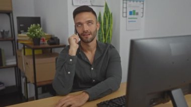 Genç bir İspanyol adam, profesyonel ve modern bir iç mekan ortamını vurgulayarak düzenli bir ofis çalışma alanında masasında telefonuyla konuşuyor..
