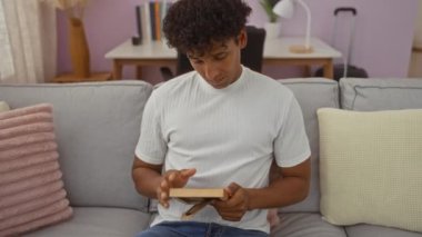 Genç bir adam rahat bir oturma odasında bir kanepede otururken fotoğraf çerçevesi tutuyor, arkada modern mobilyalar ve yumuşak yastıklarla duygularını ve nostaljisini ifade ediyor..