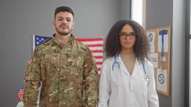 Kendine güvenen bir kadın doktor ve bir erkek asker, Amerikan bayrağıyla süslenmiş bir sağlık merkezinde çapraz kollarla dururlar..