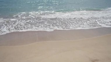 Güneşli bir gökyüzünün altında kumlu sahilde hafif dalgalarla dalgalanan sakin bir sahil.