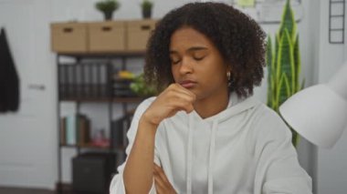 Ofis ortamında kıvırcık saçlı Afro-Amerikalı bir kadın masada otururken düşünceli ve düşünceli görünüyor..