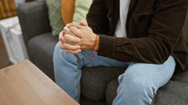 Bileziği olan genç bir adam evde oturur. Kot pantolon ve elleri bağlı bir kazak giyer..