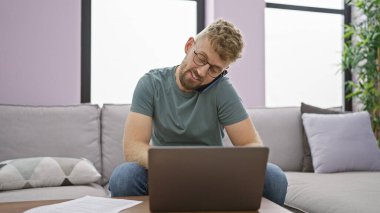 Yakışıklı, sakallı, gözlüklü, dizüstü bilgisayar kullanan ve oturma odasında telefonla konuşan beyaz adam.