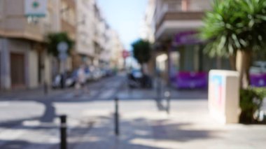 Defocused street scene with blurry background of urban outdoor area featuring buildings, cars, and a person walking in bright daylight clipart