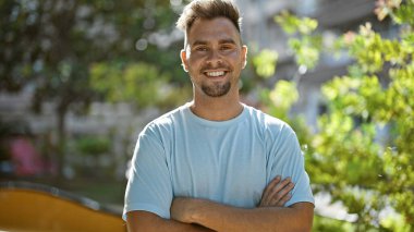 Gülümseyen, sakallı, sıradan giyinmiş, güneşli bir parkta kolları çapraz duran İspanyol bir adam..