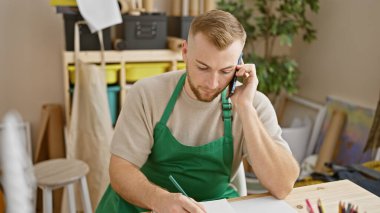 Yaratıcı bir atölyede önlük giyen sakallı genç bir adam telefonda konuşur ve not alır.