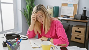 Ofis masasında başı, evrakları ve telefonu olan pembe tişörtlü stresli sarışın kadın..