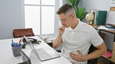 Hispanik adam modern ofiste kendini kötü hissediyor, dizüstü bilgisayar ve kalemlerin yanında karnını sıkıyor.