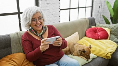Köpeğiyle akıllı telefon kullanan, rahat oturma odasında kanepede oturan gülümseyen olgun bir kadın..