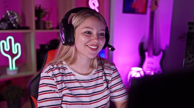 Genç sarışın bir kadın, gecenin bir yarısı canlı bir ev odasında, kulaklık takarak oyun oynuyor..