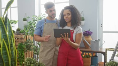 Çiçekçi dükkanında çiçeklerin arasında tablet kullanan önlüklü kadın ve erkek.