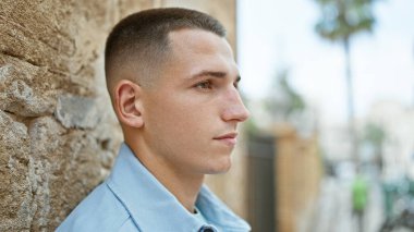 Güneşli bir şehir ortamında düşünceli bir şekilde poz veren, günlük kıyafetli yakışıklı İspanyol bir adam.