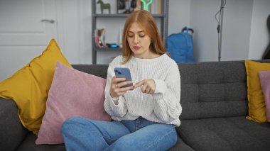 Güzel bir genç kadın, modern bir oturma odasındaki rahat rahat bir koltukta dinlenirken akıllı telefon kullanır..