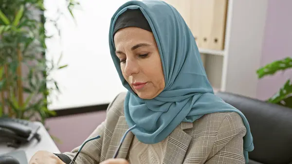 Stock image Mature woman wearing hijab focused on work in a modern office, holding glasses, portrays diversity and inclusion in the workplace.