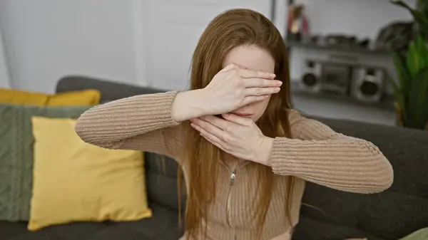 stock image A young woman covering her face with hands sitting on a sofa indoors, portraying stress or sadness emotionally.