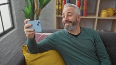 Yetişkin sakallı bir adam telefonla selfie çekiyor, kitap rafı arka planında rahat bir oturma odasında gülümsüyor..