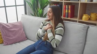 İç açıcı bir portre, genç, gülümseyen İspanyol bir kadın evdeki kanepede oturuyor, minnettarlığını gösteriyor, elleri göğsünde sağlıklı bir mutluluk içinde. Dost canlısı, neşeli oturma odası atmosferi..