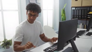 Ofiste çalışan genç bir adam, bir masaüstü bilgisayarı ve akıllı telefonu kullanarak, fabrikaları, pencereleri ve organize rafları olan modern bir ofis ortamında çalışıyor.