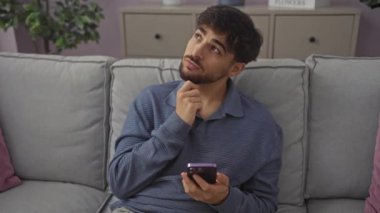 Düşünceli genç Arap adam, evinde koltukta oturuyor, eli çenesinde, bir fikri inceliyor, yüzünde ciddi bir ifade, düşünceleri sorguluyor, akıllı telefon kullanıyor.