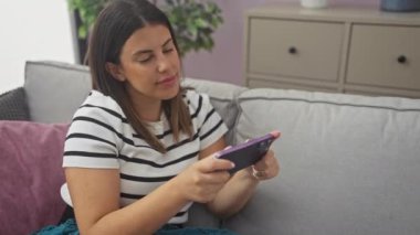 Kanepede oturan çizgili tişörtlü genç İspanyol kadın evinin oturma odasında akıllı telefonuyla oynuyor..