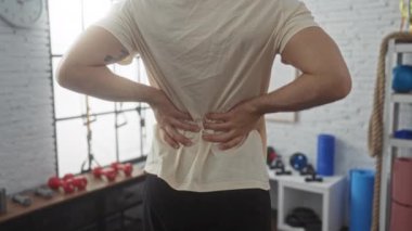 Sırt ağrısı çeken bir adam arka planda çeşitli fitness ekipmanları olan kapalı bir spor salonunda iki eliyle belini tutar..