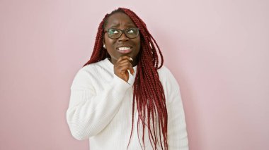 Gözlük takan ve pembe arka planda beyaz kazak giyen düşünceli Afro-Amerikan bir kadın.