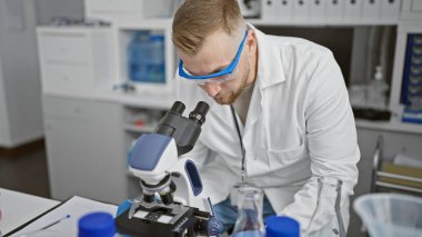 Laboratuvarda konsantre olmuş genç bir adam koruyucu gözlük takmış mikroskopla numuneyi inceliyor..