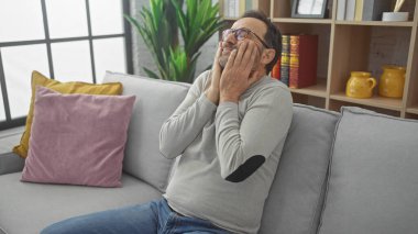Stresli, orta yaşlı, sakallı bir adam gri bir koltukta oturuyor. Endişe ya da tefekkür belirtileri gösteriyor.