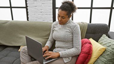 Kıvırcık saçlı genç bir kadın rahat bir oturma odasında dizüstü bilgisayarla çalışıyor..