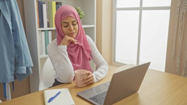 Kafiyeli sakin bir kadın ev ofisinde dizüstü bilgisayar kullanırken kahveyi yudumluyor, modern ve profesyonel hayatı yansıtıyor..