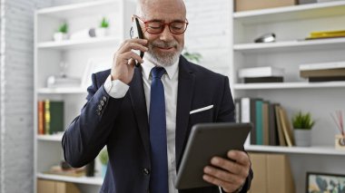 Sakallı olgun bir adam, modern bir ofis ortamında elinde tabletle telefon görüşmesi yapar..