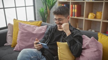 Neşeli, genç, İspanyol bir adam modern bir oturma odasında renkli bir kanepede akıllı telefonuyla dinleniyor..