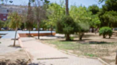 Bokeh etkisinden arındırılmış açık hava sahnesi bulanık bir park yolu, yeşil ağaçlar ve açık bir gökyüzü altında uzak binalar, canlı ve huzurlu bir atmosfer yaratıyor..